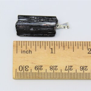 May include: A black tourmaline pendant with a silver-colored bail. The raw, natural crystal is approximately 2 inches long. The pendant is displayed on a white surface next to a wooden ruler marked in inches.