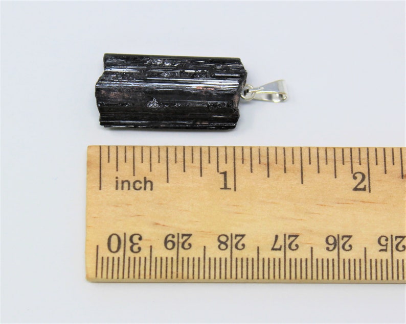 May include: A black tourmaline pendant with a silver-colored bail. The raw, natural crystal is approximately 2 inches long. The pendant is displayed on a white surface next to a wooden ruler marked in inches.