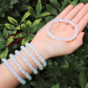 May include: A set of six white opal beaded bracelets stacked on a person's wrist. The bracelets are made of round, smooth beads.