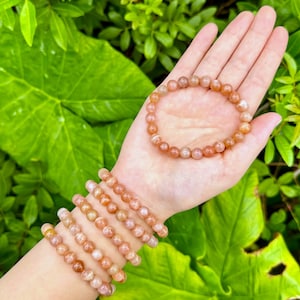 May include: A set of six sunstone bracelets with orange and pink beads. The bracelets are stacked on a person's wrist.