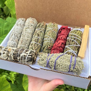 May include: A box of assorted smudge sticks, including white sage, cedar, palo santo, and lavender. The sticks are bundled together with twine and are ready to be used for cleansing and purification rituals.