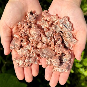 May include: A collection of raw copper nuggets with a metallic, reddish-brown luster and irregular, jagged shapes. The pieces exhibit a textured, porous surface with varying shades of copper, ranging from bright metallic tones to darker oxidized areas.