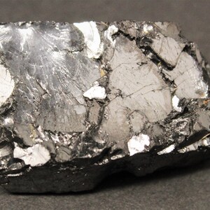 May include: A close-up of a rough, dark gray mineral specimen with a metallic sheen. The surface is fractured, revealing a mix of dark gray and silver-white crystalline structures. The overall shape is rectangular.