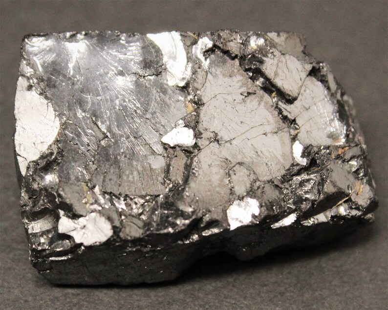 May include: A close-up of a rough, dark gray mineral specimen with a metallic sheen. The surface is fractured, revealing a mix of dark gray and silver-white crystalline structures. The overall shape is rectangular.