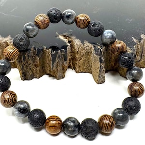 May include: A beaded bracelet featuring a mix of natural stone beads. The bracelet includes dark gray lava rock beads, gray-blue stone beads, and brown wooden beads. The beads are strung together to form a circular shape.