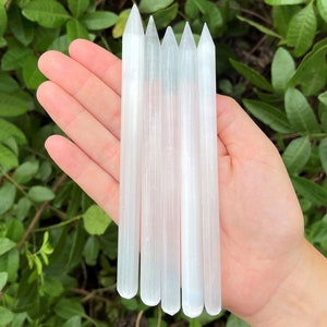 May include: Six white selenite wands, also known as selenite sticks, are held in a hand. The wands are smooth and have a natural, striated texture.