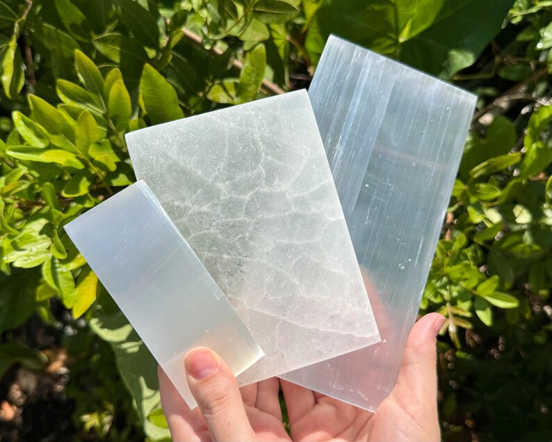 May include: Three rectangular selenite slabs of varying sizes. One slab is translucent with a smooth surface, one is opaque with a white crackled texture, and the third is a long, clear, fibrous piece with a distinct linear grain pattern.