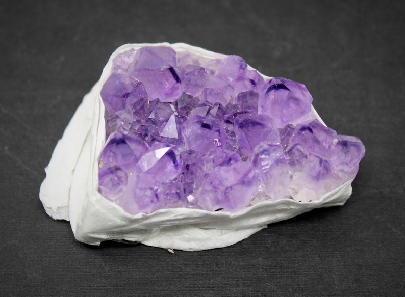 May include: A cluster of amethyst crystals in shades of purple, set in a white, natural matrix. The crystals are arranged in a natural formation, showcasing their unique shapes and facets.