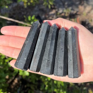 May include: Six black obsidian points, also known as black glass, are arranged in a row on a hand. The points are smooth and polished, with a slight sheen.
