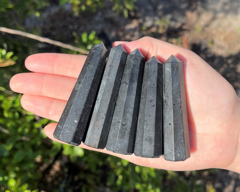 May include: Six black obsidian points, also known as black glass, are arranged in a row on a hand. The points are smooth and polished, with a slight sheen.