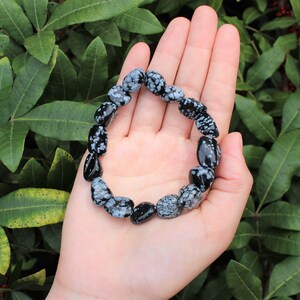 May include: A black and white snowflake obsidian bracelet made of tumbled stones. The bracelet is on a hand, with green foliage in the background.