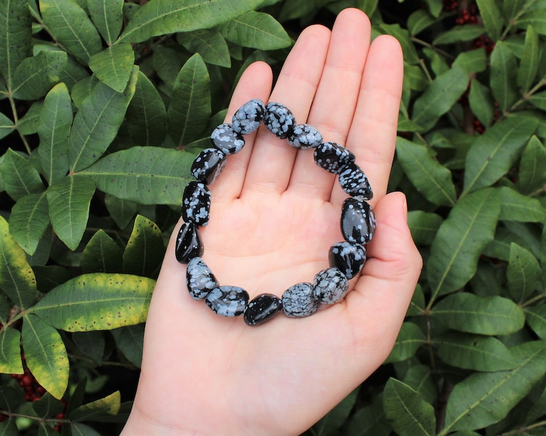 May include: A black and white snowflake obsidian bracelet made of tumbled stones. The bracelet is on a hand, with green foliage in the background.