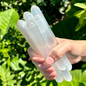 May include: A bundle of white selenite wands, also known as selenite sticks, are held in a hand. The wands are smooth and have a striated texture.