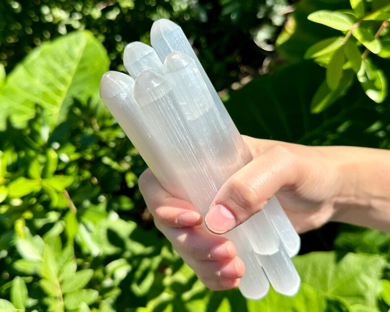 May include: A bundle of white selenite wands, also known as selenite sticks, are held in a hand. The wands are smooth and have a striated texture.