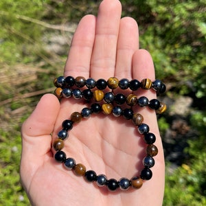 Pode incluir: Um bracelete feito de contas de hematita preta, olho de tigre e &ocirc;nix preto. O bracelete &eacute; mostrado em uma m&atilde;o contra um fundo verde.