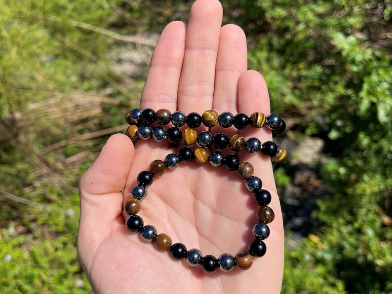 Pode incluir: Um bracelete feito de contas de hematita preta, olho de tigre e &ocirc;nix preto. O bracelete &eacute; mostrado em uma m&atilde;o contra um fundo verde.