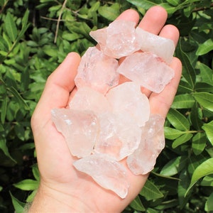 May include: A collection of raw, clear quartz crystals with a slight pink hue. The crystals are various sizes and shapes, and they are arranged in a pile in the palm of a hand.