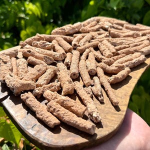 May include: A wooden serving board holds a collection of tan, cylindrical objects. The objects have a textured, rough appearance and are arranged in a pile. The board is a warm brown color, and the background is blurred green foliage.