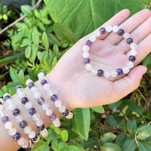 May include: A set of four bracelets made with amethyst, rose quartz, and clear quartz beads. The bracelets are stacked on a person's wrist.