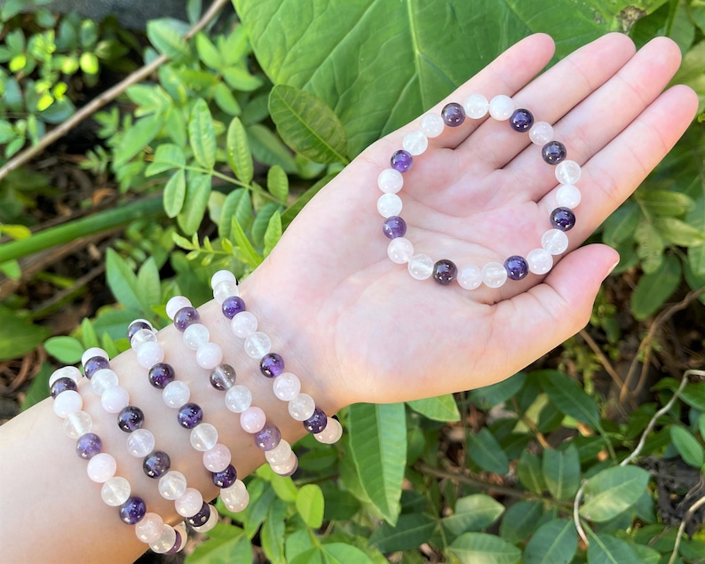 May include: A set of four bracelets made with amethyst, rose quartz, and clear quartz beads. The bracelets are stacked on a person's wrist.