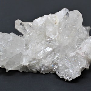 May include: A cluster of clear quartz crystals with a variety of sizes and shapes. The crystals are arranged in a natural, organic pattern, and some have a slight iridescent sheen.