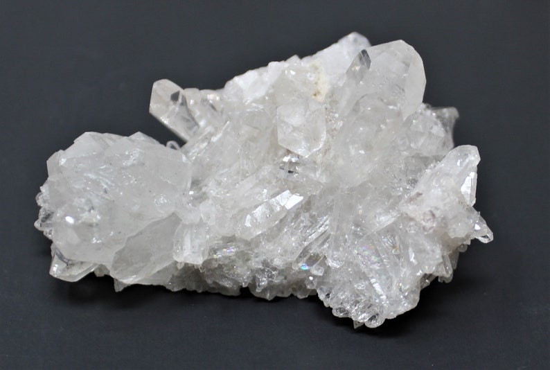 May include: A cluster of clear quartz crystals with a variety of sizes and shapes. The crystals are arranged in a natural, organic pattern, and some have a slight iridescent sheen.