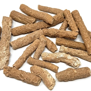 May include: A collection of brown, stick-shaped treats, possibly dog chews, scattered on a white surface. The treats vary in size and texture, with a rough, granular appearance. The color ranges from light tan to a darker brown.