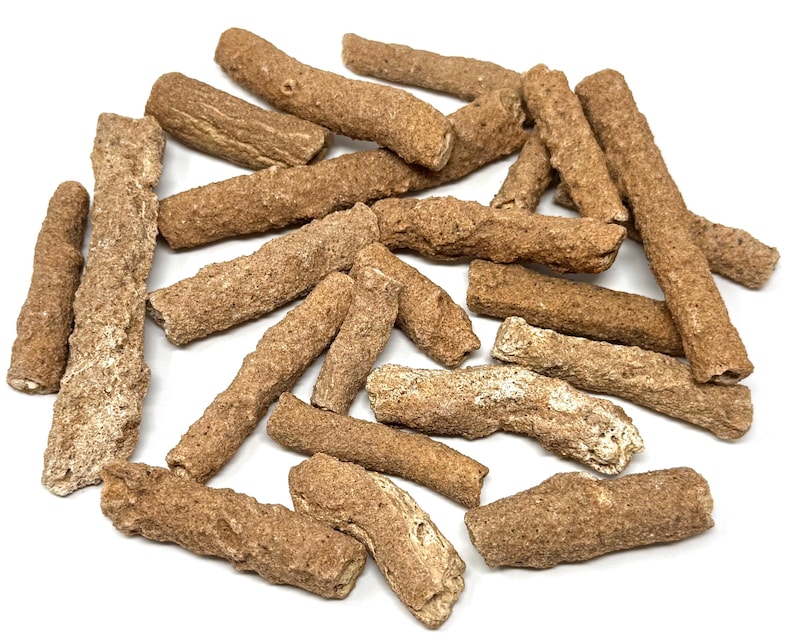 May include: A collection of brown, stick-shaped treats, possibly dog chews, scattered on a white surface. The treats vary in size and texture, with a rough, granular appearance. The color ranges from light tan to a darker brown.