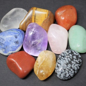 May include: A collection of ten tumbled stones in various colors, including clear, blue, purple, pink, green, brown, red, and orange. The stones are arranged in a circle on a dark gray surface.