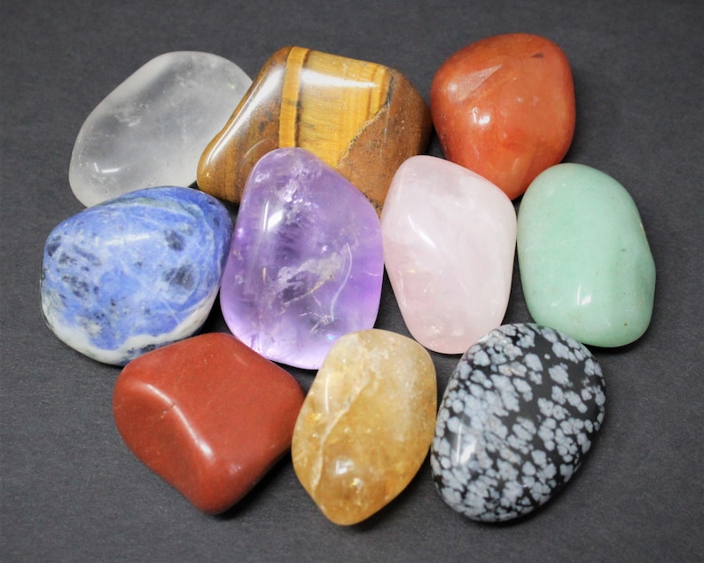 May include: A collection of ten tumbled stones in various colors, including clear, blue, purple, pink, green, brown, red, and orange. The stones are arranged in a circle on a dark gray surface.