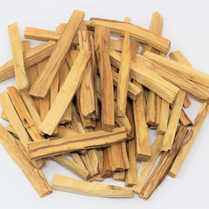 May include: A collection of light brown Palo Santo wood sticks, showing natural wood grain and diverse shapes. The rectangular sticks look freshly cut, with a natural, earthy appearance. The sticks are scattered on a white surface.