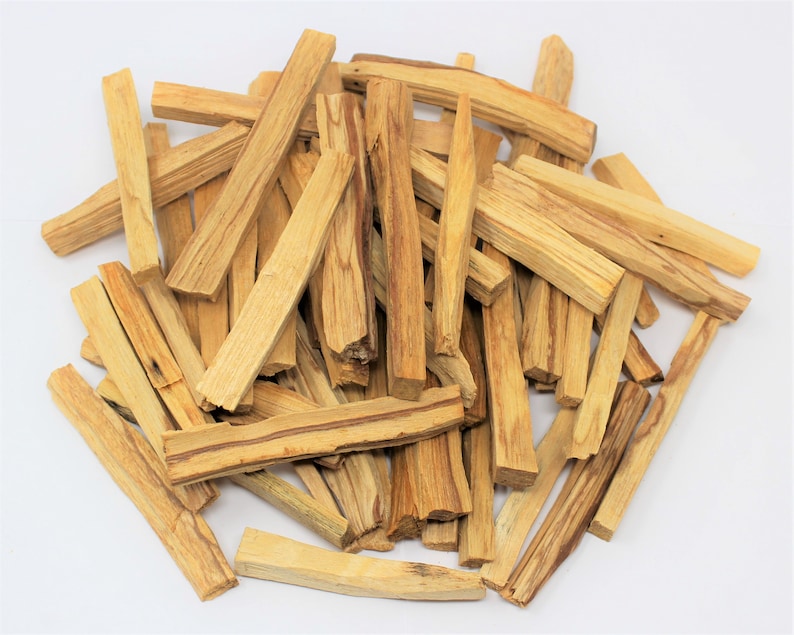 May include: A collection of light brown Palo Santo wood sticks, showing natural wood grain and diverse shapes. The rectangular sticks look freshly cut, with a natural, earthy appearance. The sticks are scattered on a white surface.