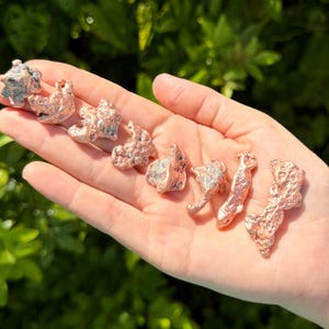 May include: A collection of eight small, irregular copper-colored metal nuggets with a textured, porous surface. The pieces vary in shape and size, displaying a metallic sheen and a reddish-brown hue, arranged in a line across the palm of a hand.