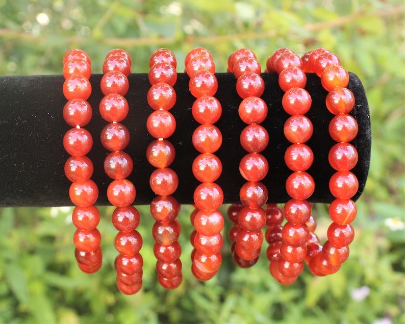 May include: Several bracelets with large, round, orange-red beads are displayed against a black background. The beads are strung together, creating a continuous loop. The bracelets are likely made of a gemstone or similar material.