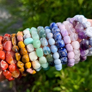 May include: A collection of multiple stretch bracelets featuring polished, tumbled gemstone beads in a spectrum of colors including red, orange, yellow, green, blue, purple, and white, arranged in a row on a wrist against a blurred green background.