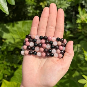 May include: A stack of four bracelets made with pink, white, and black gemstone beads. The bracelets are made with round beads and are stacked on top of each other.
