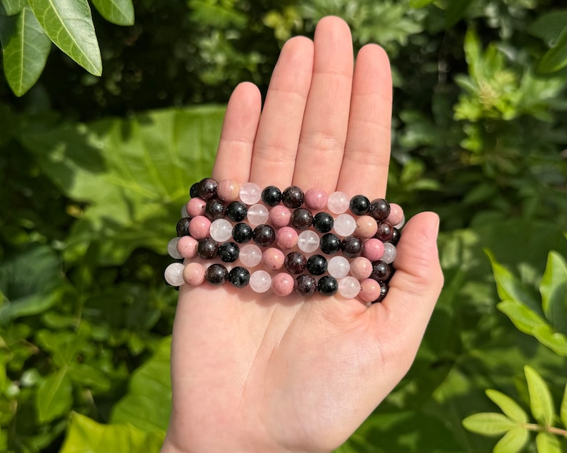 May include: A stack of four bracelets made with pink, white, and black gemstone beads. The bracelets are made with round beads and are stacked on top of each other.