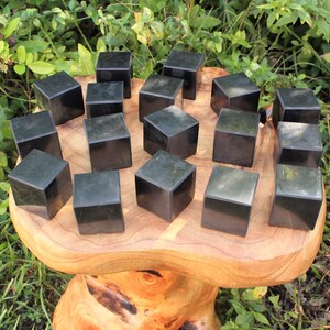 May include: A collection of 18 black obsidian cubes arranged in a circular pattern on a wooden surface. The cubes are smooth and polished, with a slight sheen.