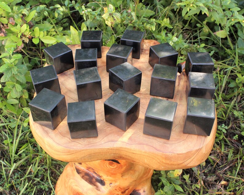 May include: A collection of 18 black obsidian cubes arranged in a circular pattern on a wooden surface. The cubes are smooth and polished, with a slight sheen.