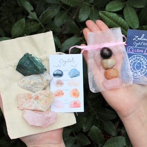 May include: Two small bags containing crystals. The bag on the left is made of beige fabric and contains a variety of crystals, including green, white, orange, and pink. The bag on the right is made of pink tulle and contains three crystals: a dark brown crystal, a light brown crystal, and a pink crystal. A card with the text "Zodiac Crystal Kit Libra" is visible in the image.