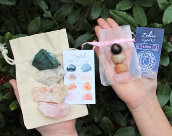 Pode incluir: Duas pequenas bolsas contendo cristais. A bolsa à esquerda é feita de tecido bege e contém uma variedade de cristais, incluindo verde, branco, laranja e rosa. A bolsa à direita é feita de tule rosa e contém três cristais: um cristal marrom escuro, um cristal marrom claro e um cristal rosa. Um cartão com o texto "Zodiac Crystal Kit Libra" é visível na imagem.