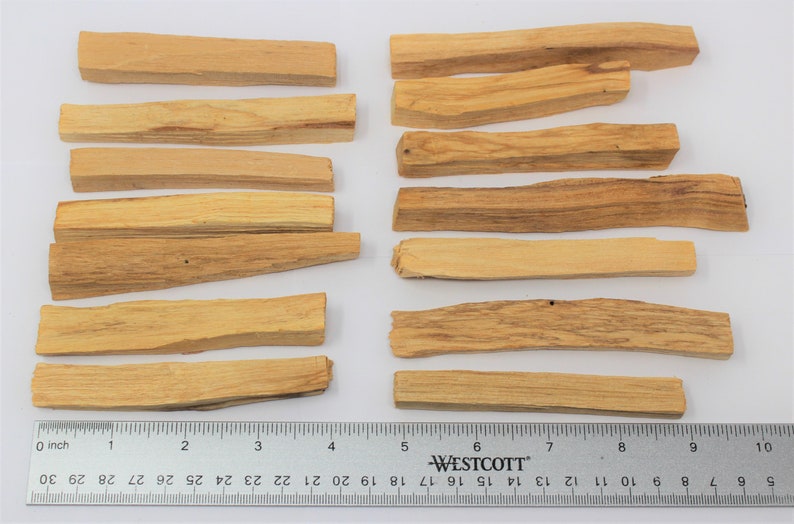 May include: Several rectangular Palo Santo wood sticks, light brown in colour, are arranged on a white surface. A ruler with inch measurements is placed below the sticks. The wood pieces have a natural, textured appearance.