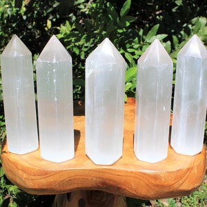 May include: Five translucent, white selenite crystal towers with pointed tops are displayed on a wooden stand. The crystals are arranged in a row, each with a rectangular base and a faceted, pyramid-shaped top. The background features green foliage.