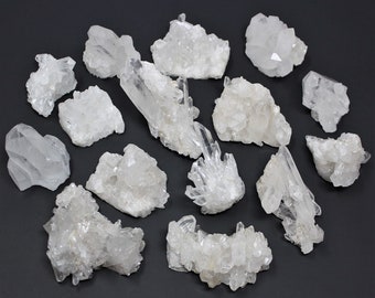 May include: A collection of 15 clear quartz crystal clusters of varying sizes and shapes. The crystals are arranged on a dark grey surface.
