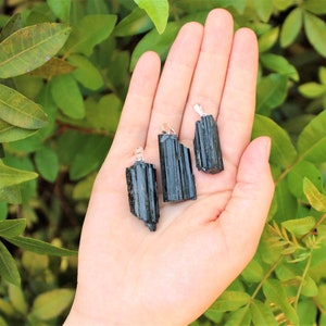 May include: Three black tourmaline pendants with silver-colored bails are displayed against a backdrop of green foliage. The raw, natural crystals vary slightly in size and shape, showcasing their unique textures and dark color.