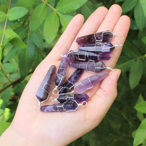 May include: A collection of amethyst crystal points wrapped in silver wire, each with a loop for hanging. The crystals are various shades of purple and are displayed on a hand.