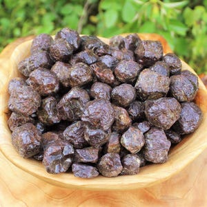 May include: A collection of raw, dark reddish-brown garnet stones piled inside a light-colored wooden bowl. The stones have an irregular, multifaceted, and textured surface with a natural luster, filling the bowl to the brim with various sizes.