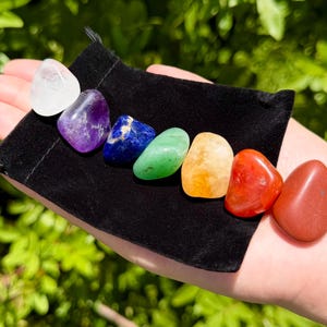 May include: A set of seven polished, tumbled gemstones arranged in a row on a black velvet drawstring pouch. The stones include clear quartz, amethyst, sodalite, green aventurine, citrine, carnelian, and red jasper in a spectrum of colors.