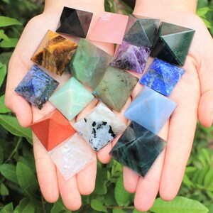 Puede incluir: Una colecci&oacute;n de varias pir&aacute;mides de piedras preciosas de colores, incluyendo negro, rosa, verde, morado, azul y rojo. Las pir&aacute;mides se sostienen en manos abiertas sobre un fondo de follaje verde. Cada pir&aacute;mide tiene un color y patr&oacute;n &uacute;nicos.