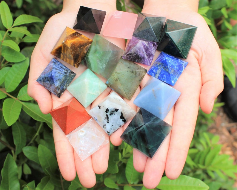 Puede incluir: Una colecci&oacute;n de varias pir&aacute;mides de piedras preciosas de colores, incluyendo negro, rosa, verde, morado, azul y rojo. Las pir&aacute;mides se sostienen en manos abiertas sobre un fondo de follaje verde. Cada pir&aacute;mide tiene un color y patr&oacute;n &uacute;nicos.